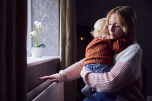 Mother carrying child in home whilst holding one hand over the radiator for warmth.