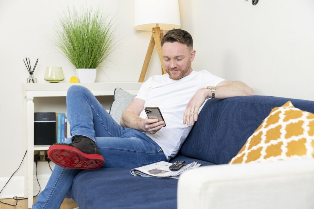 A man in white shirt and blue jeans on a sofa, reading an energy advice email on his phone