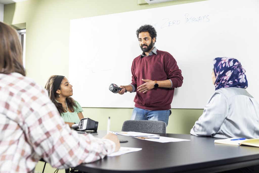 3 people round a table, watching a man in front of a whiteboard